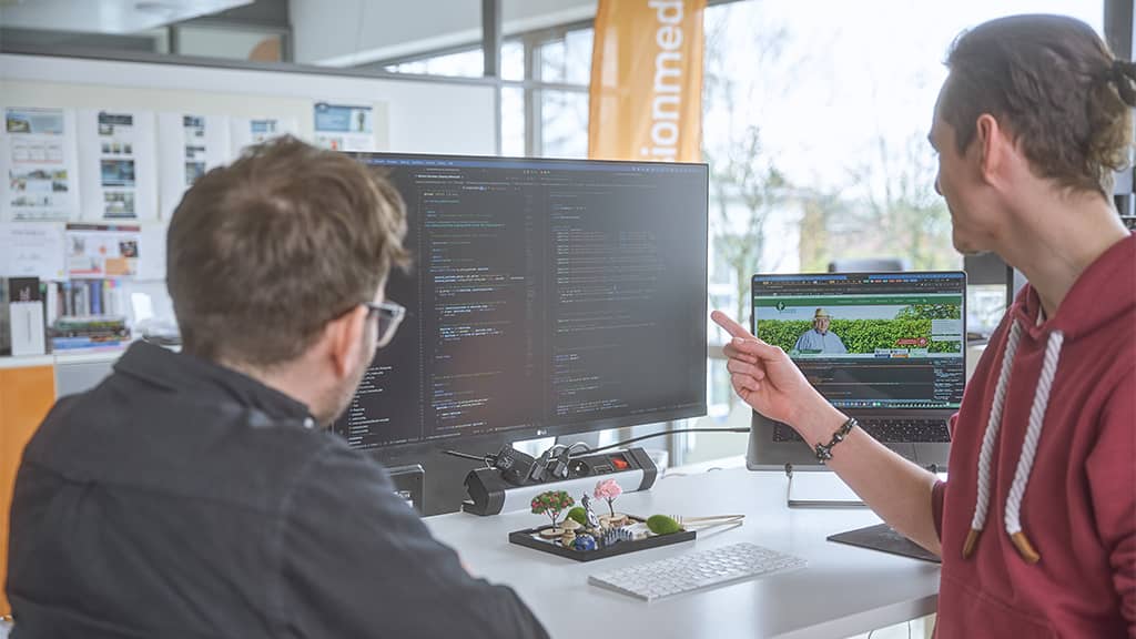 Zwei Entwickler vergleichen Code auf einem großen Monitor und eine geöffnete Webseite auf einem Laptop im Büro, Fokus auf barrierefreie Webentwicklung.