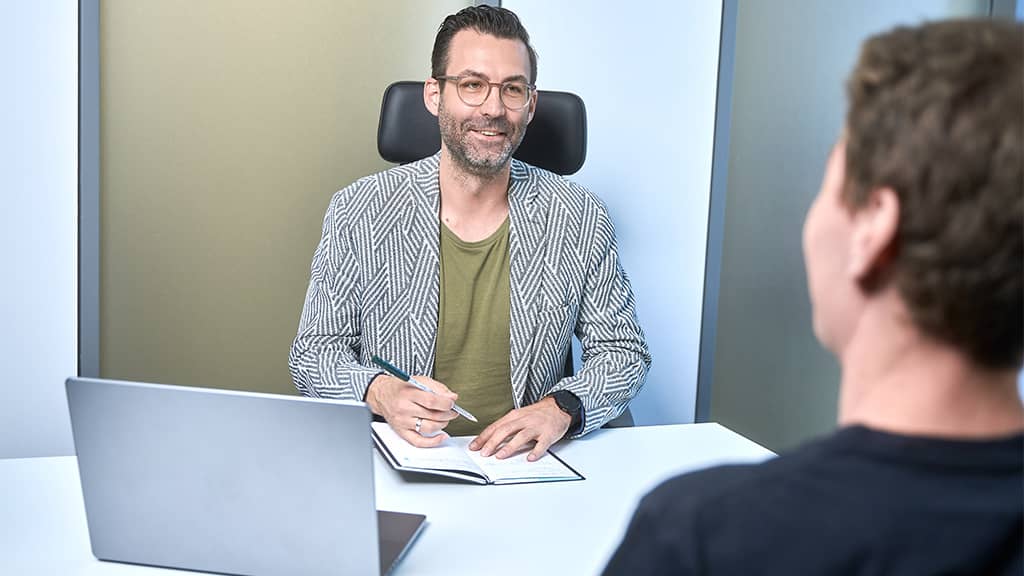 Berater sitzt lächelnd am Schreibtisch, macht Notizen und spricht mit einem Klienten; Laptop auf dem Tisch.