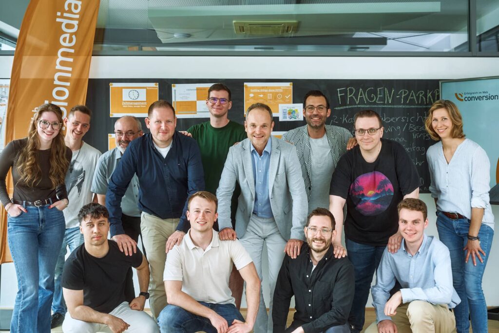 Das Conversionmedia-Team posiert lächelnd gemeinsam im Büro vor einer Präsentationswand.