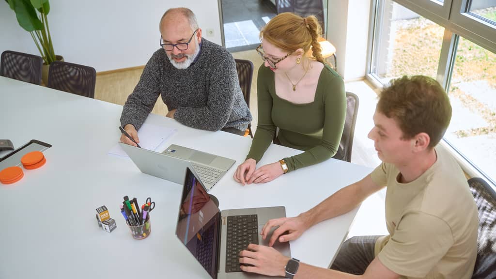 Älterer Mann, junge Frau und junger Mann sitzen an einem Tisch und nutzen Laptops, Notizen und Stifte liegen bereit.