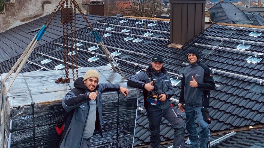 Drei GEBA-Mitarbeiter geben Daumen hoch auf einem Dach, während Material per Kran positioniert wird.