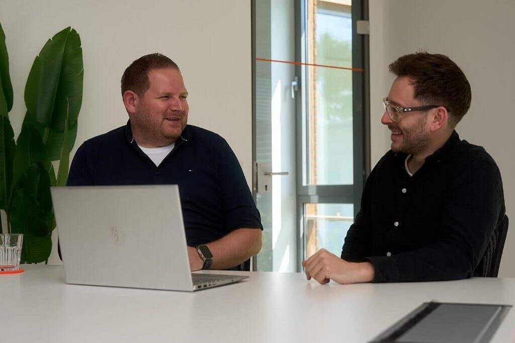 Zwei Männer unterhalten sich entspannt an einem Besprechungstisch mit offenem Laptop.