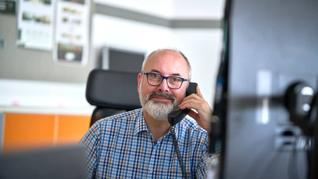 Ein Mann mit kariertem Hemd und Brille sitzt am Schreibtisch und spricht in ein Telefon.
