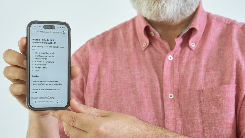 Mann in rosafarbenem Kurzarmhemd hält ein Smartphone mit lesbarem Text auf dem Display, nahe an der Kamera.