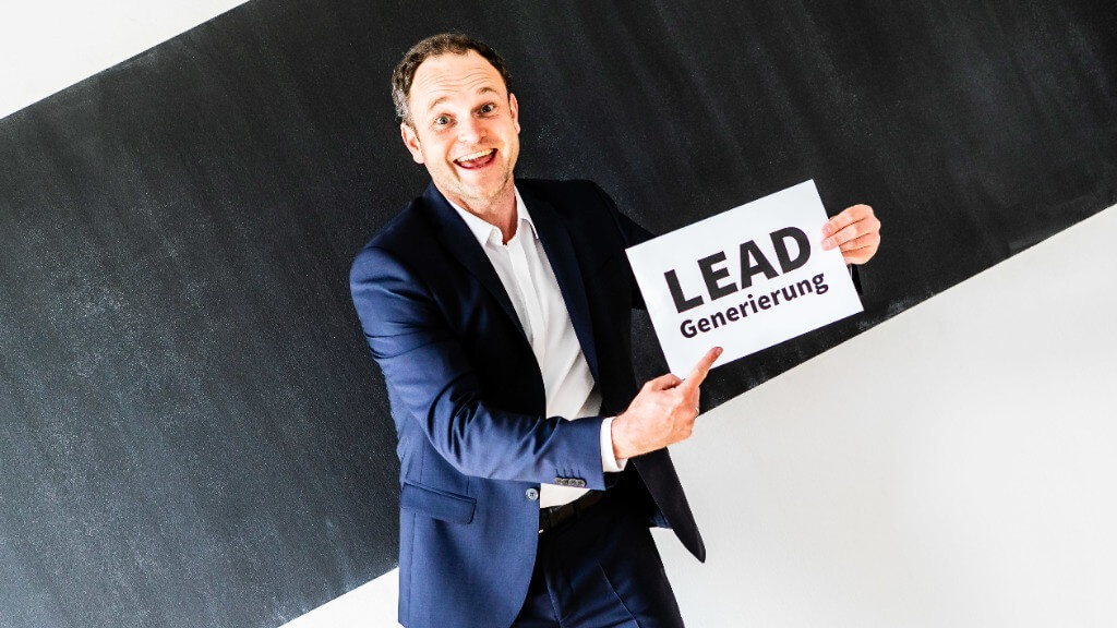 Ein lächelnder Mann im dunkelblauen Anzug hält ein weißes Schild mit dem Text ‚LEAD Generierung‘ und zeigt darauf.