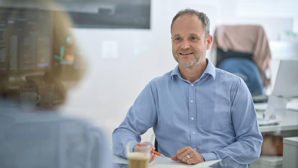 Ein lächelnder Mann in Hemd sitzt an einem Tisch, vor ihm Notizen und eine Tasse, im Hintergrund Büromöbel.