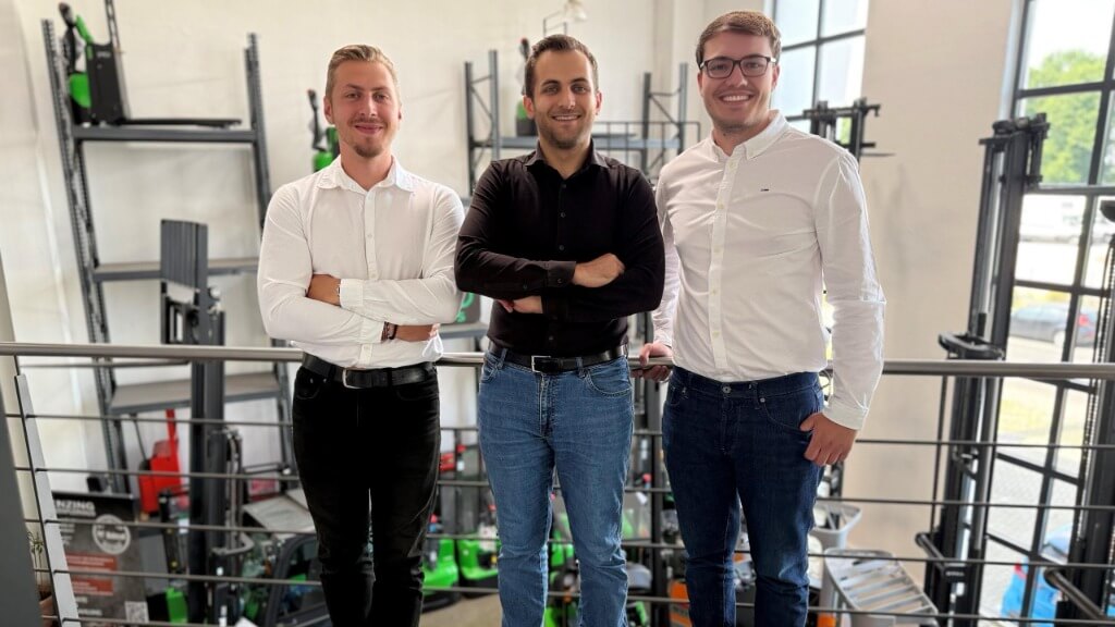 Drei Mitarbeitende des Meisterstapler-Teams stehen lächelnd in einem Lager mit Gabelstaplern im Hintergrund.
