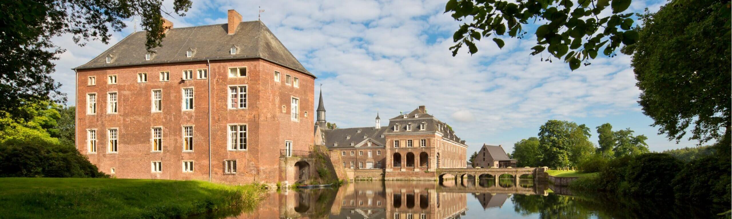 Schloss Wissen an einem klaren Tag, gespiegelt im Wassergraben mit grüner Ufervegetation.