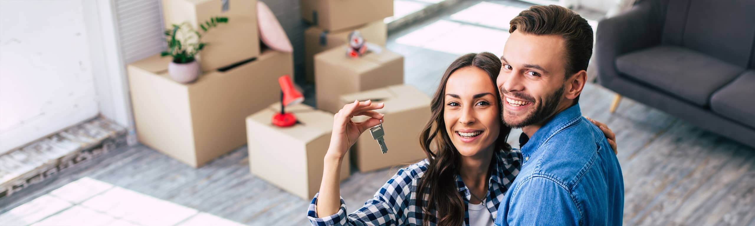 Junges Paar hält lächelnd einen Wohnungsschlüssel vor gepackten Kartons.