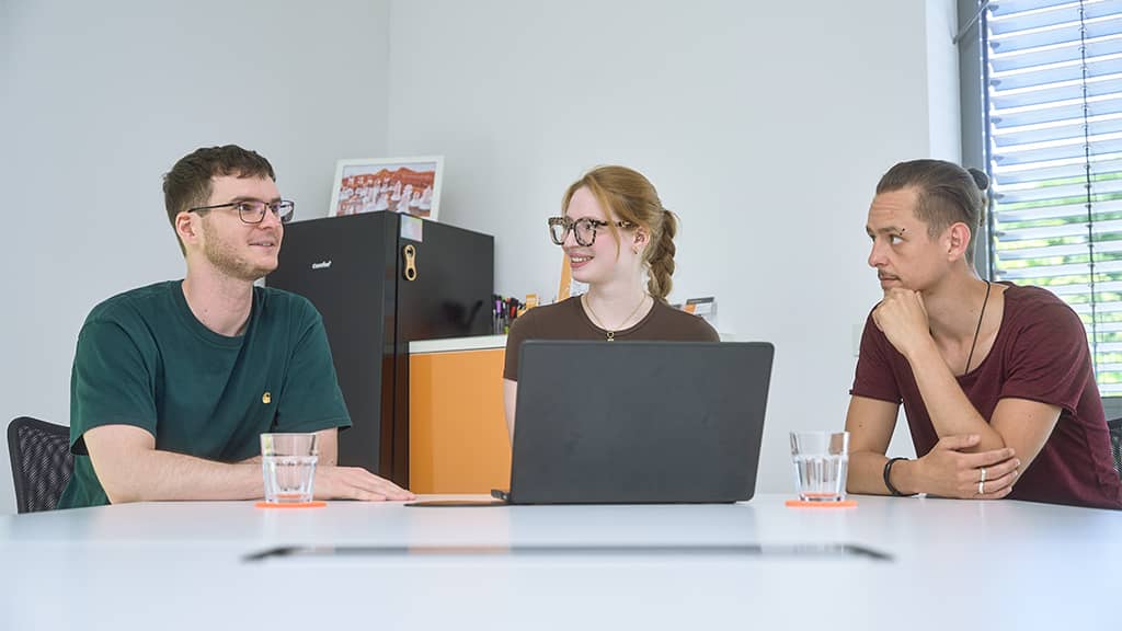Drei Personen diskutieren konzentriert an einem Tisch mit geöffnetem Laptop während eines Strategie-Workshops.