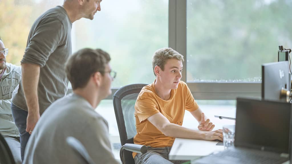 Junge Entwickler diskutieren konzentriert vor einem Bildschirm in einem modernen Büro, während ein Kollege über die Schulter blickt.