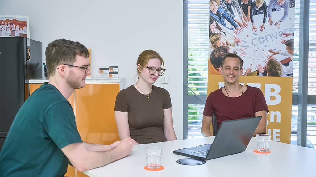 Drei junge Mitarbeitende sitzen an einem Konferenztisch und schauen gemeinsam auf einen geöffneten Laptop.