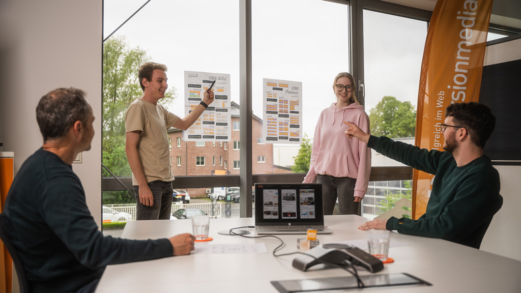 Vier Personen stehen und sitzen um einen Konferenztisch, auf dem ein Laptop steht, während zwei Poster an der Fensterscheibe befestigt sind.