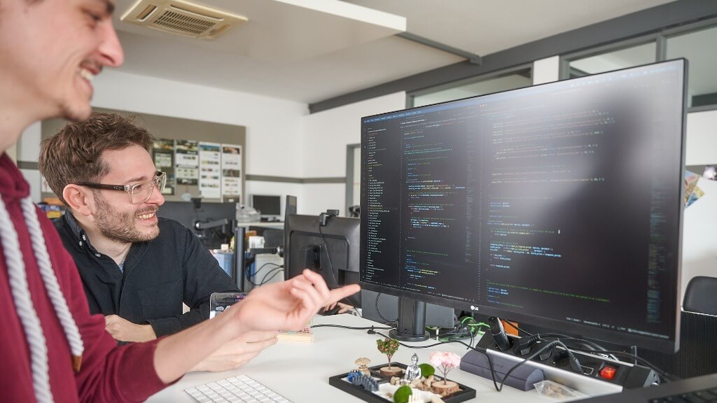 Zwei Männer sitzen an einem Schreibtisch und zeigen auf Programmcode, der auf einem großen Computerbildschirm angezeigt wird.