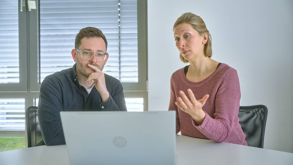 Eine Frau gestikuliert erklärend, während ein Mann nachdenklich neben ihr am Laptop sitzt.